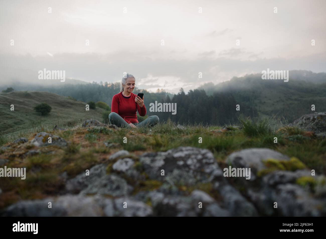 Senior woman taking selfie when doing breathing exercise in nature on early morning with fog and mountains in background. Stock Photo