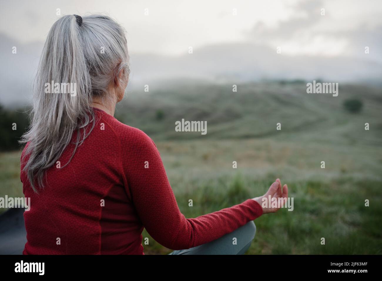 Rear view of senior woman doing breathing exercise in nature on early morning with fog and mountains in background. Stock Photo