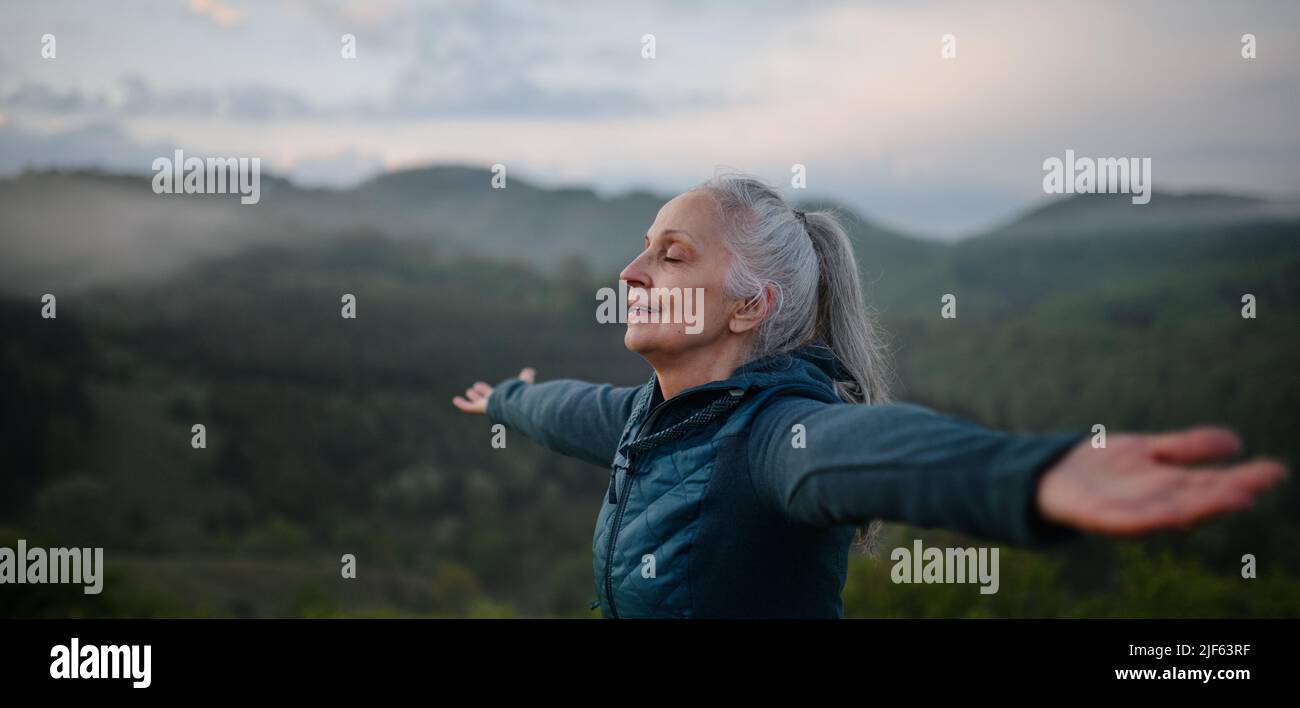 Senior woman doing breathing exercise in nature on early morning with fog and mountains in background. Stock Photo