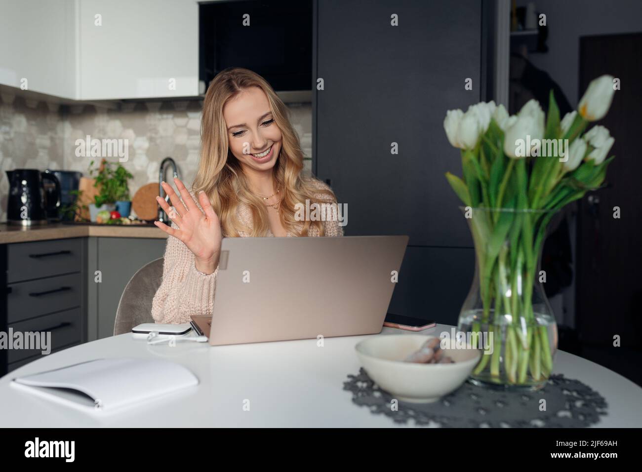 Feminine blonde in knitted jacket smiles happily, greeting her interlocutor with her hand when communicating online.  Stock Photo