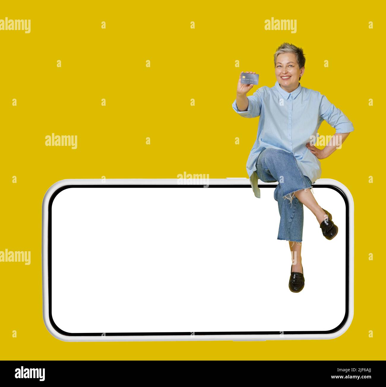 Happy grey haired mature woman sitting on a giant, huge smartphone, in casual blue shirt and jeans with hand raised up in a win, isolated on yellow background.  Stock Photo
