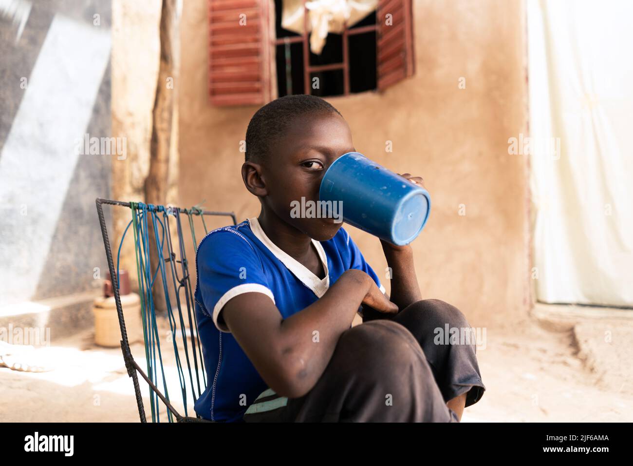 African village boy sitting in a sunny courtyard drinking from a big blue plastic mug; scarcity of water supply to private households in West African Stock Photo