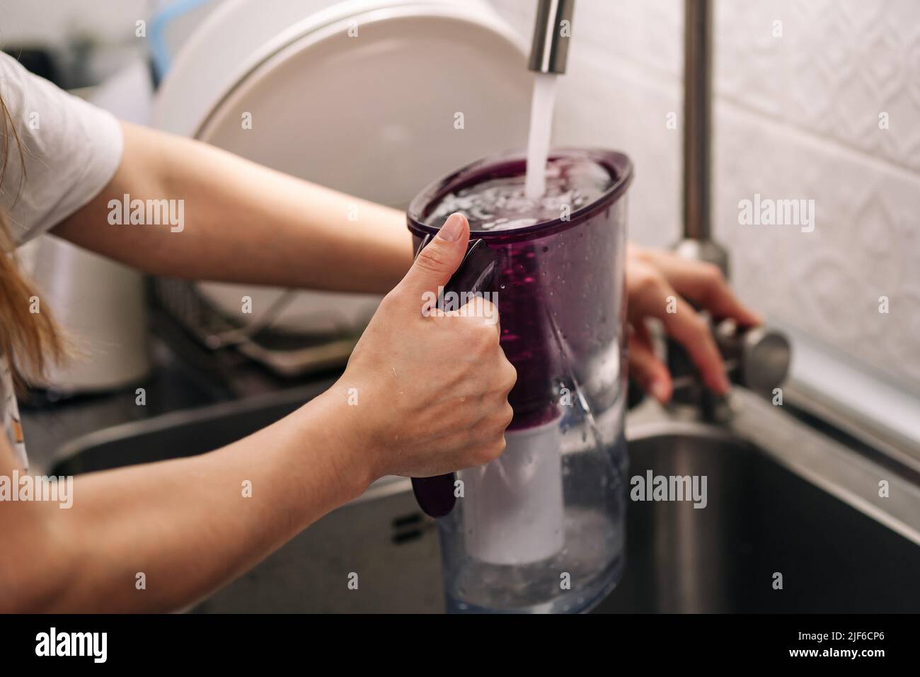 Woman pouring water from faucet into water filter jug at the kitchen. Healthy lifestyle. Woman filling water. Stock Photo