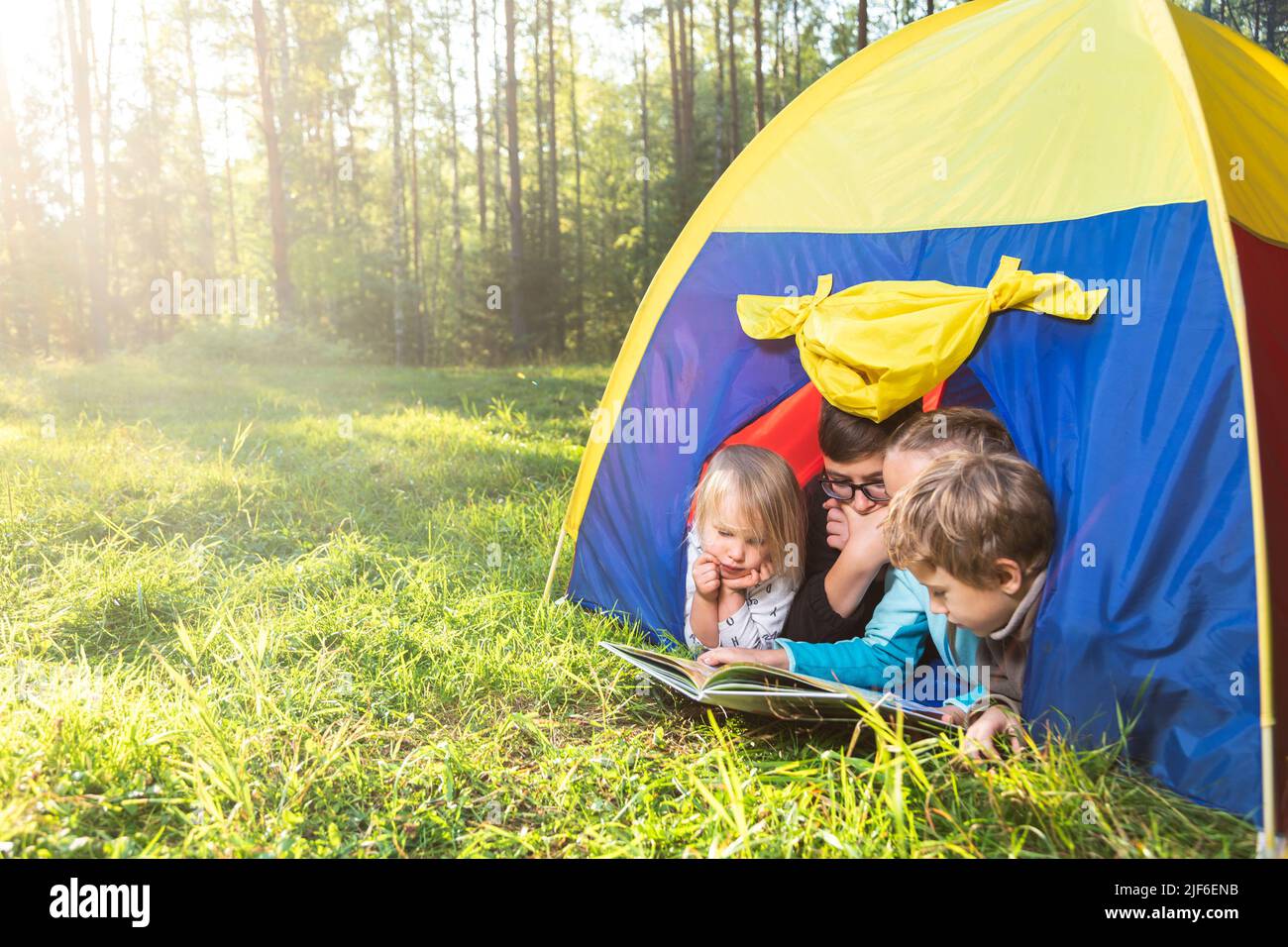 Four children of different age reading a book lying together in tent camping in a summer forest during summer holidays Stock Photo