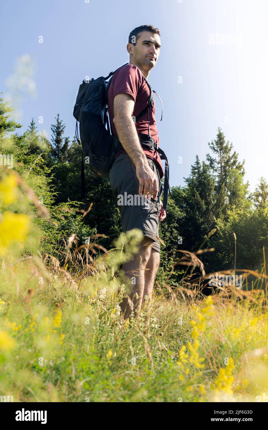 A man hiking the mountains with a backpack in the summer. Behind him is the view of the valley. He is looking at the distance behind the camera. Stock Photo