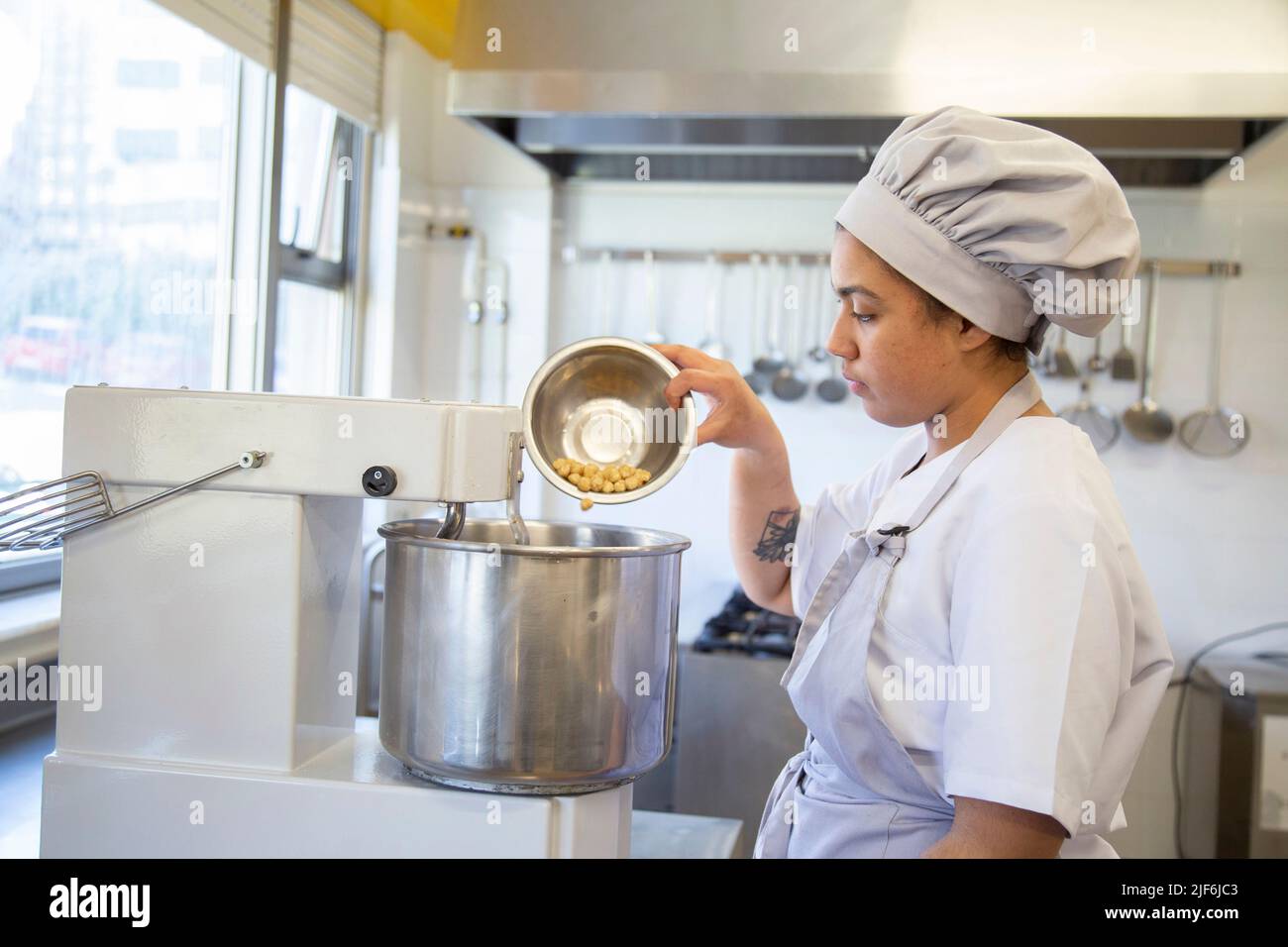 Side view of female baker in uniform pouring ingredient into metal bowl of dough mixer while standing in bakery school Stock Photo