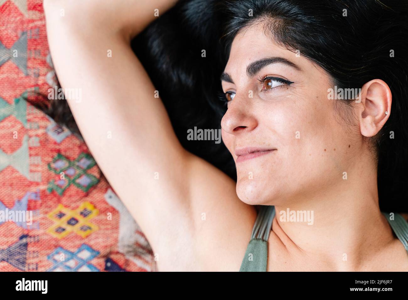 From above positive Hispanic woman with black hair and brown eyes raising arm and looking away with smile while relaxing on ornamental carpet in dayti Stock Photo