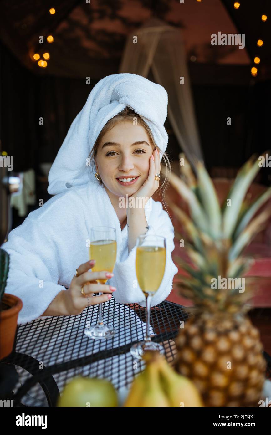 Attractive happy female wrapped in towels drinking cocktail while sitting looking at camera at table with juice and fruits on terrace of building Stock Photo