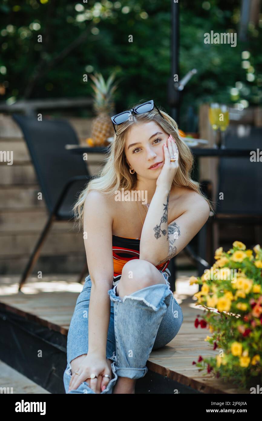 Attractive young female in casual clothes with tattoos on arm looking at camera while sitting on terrace of building on summer day Stock Photo