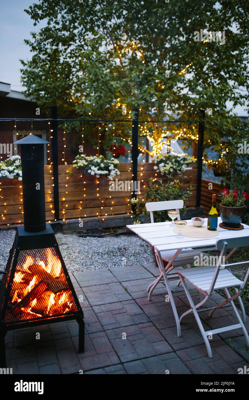 Alcohol drinks served on table near fireplace with burning firewood placed on patio near lush green trees in evening time Stock Photo