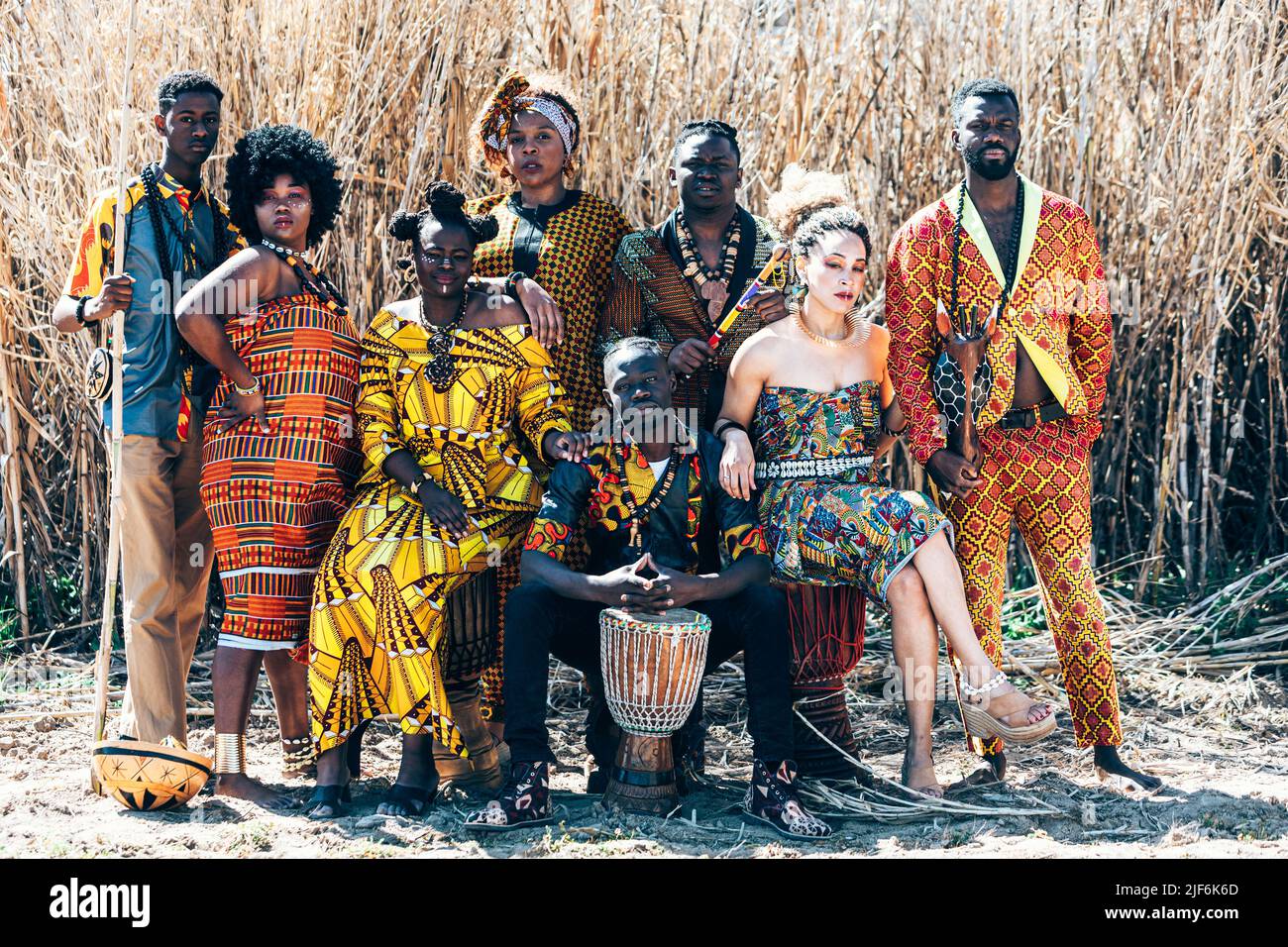 Full body group of black and mixed race women and men in colorful authentic African outfits with sticks and drums sitting and standing against dry fie Stock Photo