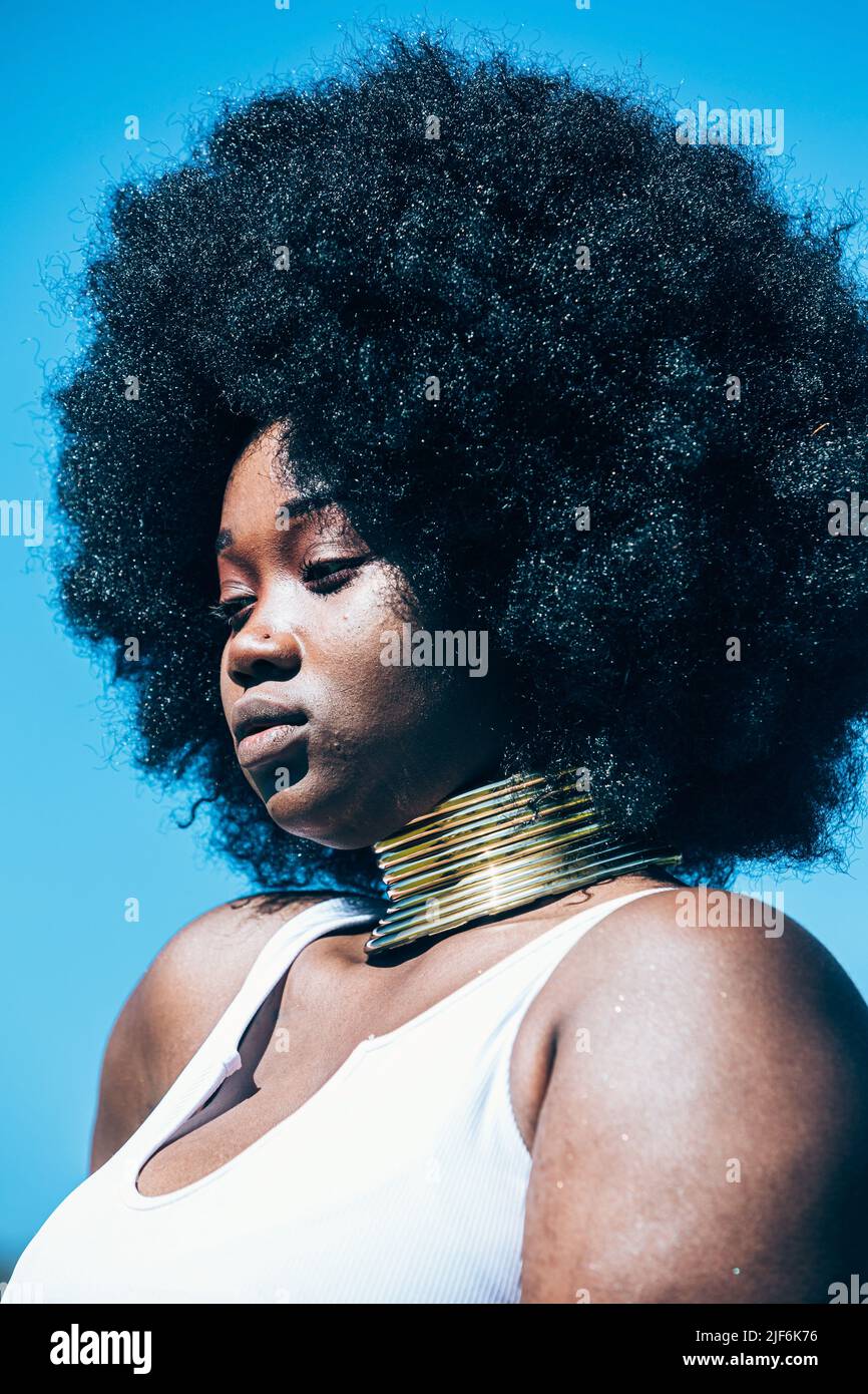 Young black woman in authentic golden necklace and Afro hairstyle looking away on blurred background of countryside Stock Photo