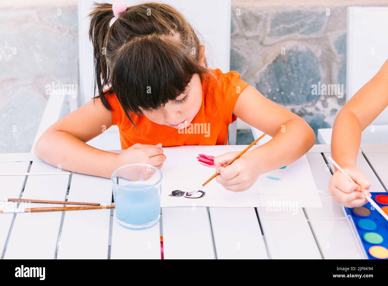Focused girl painting on paper with paintbrush at table with palette of ...