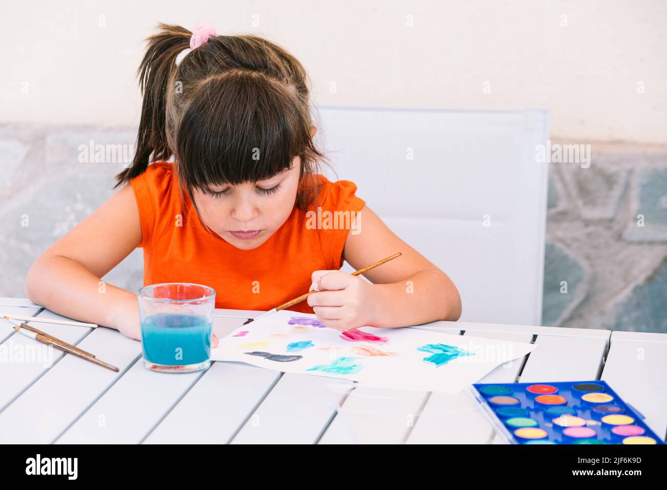 Focused girl painting on paper with paintbrush at table with palette of ...