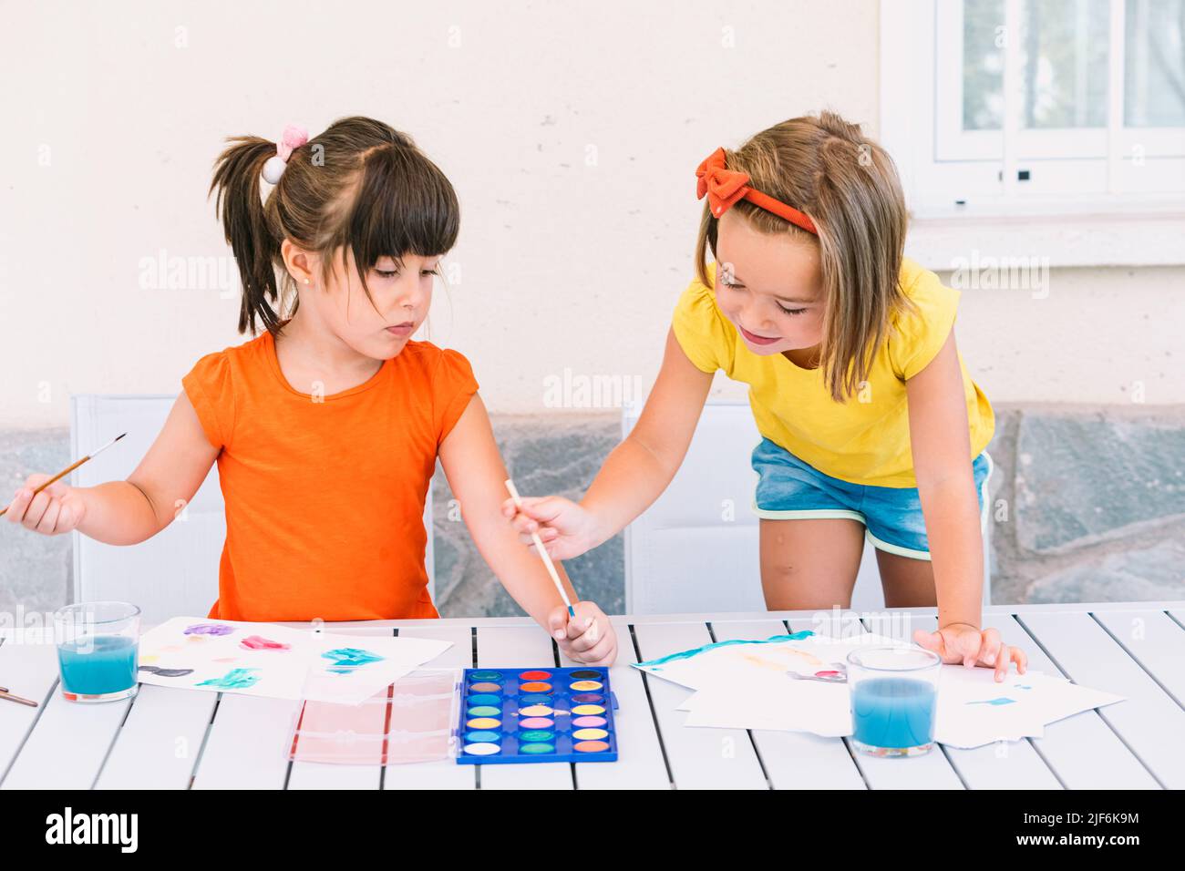 Cute girls painting with paintbrushes on papers at table with palette ...
