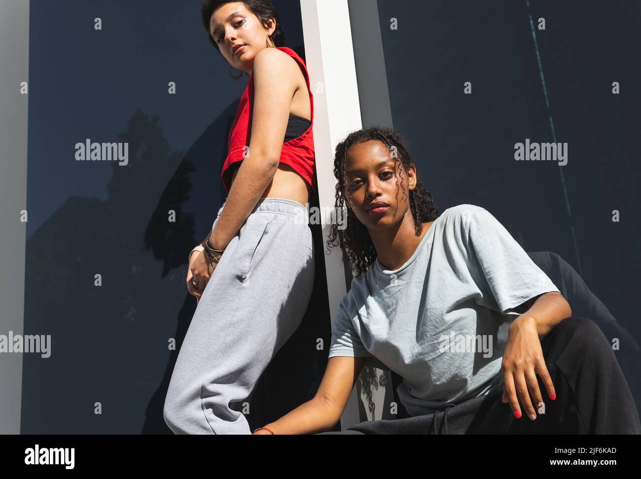 From below serious multiracial female friends in casual clothes looking at camera while standing near gray wall of building on sunny street Stock Photo