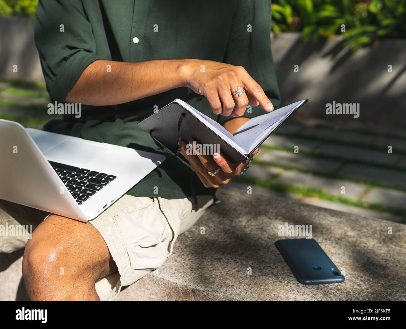 Unrecognizable male student sitting on stone bench in city park and reading notes in notepad while working on assignment using modern netbook Stock Photo