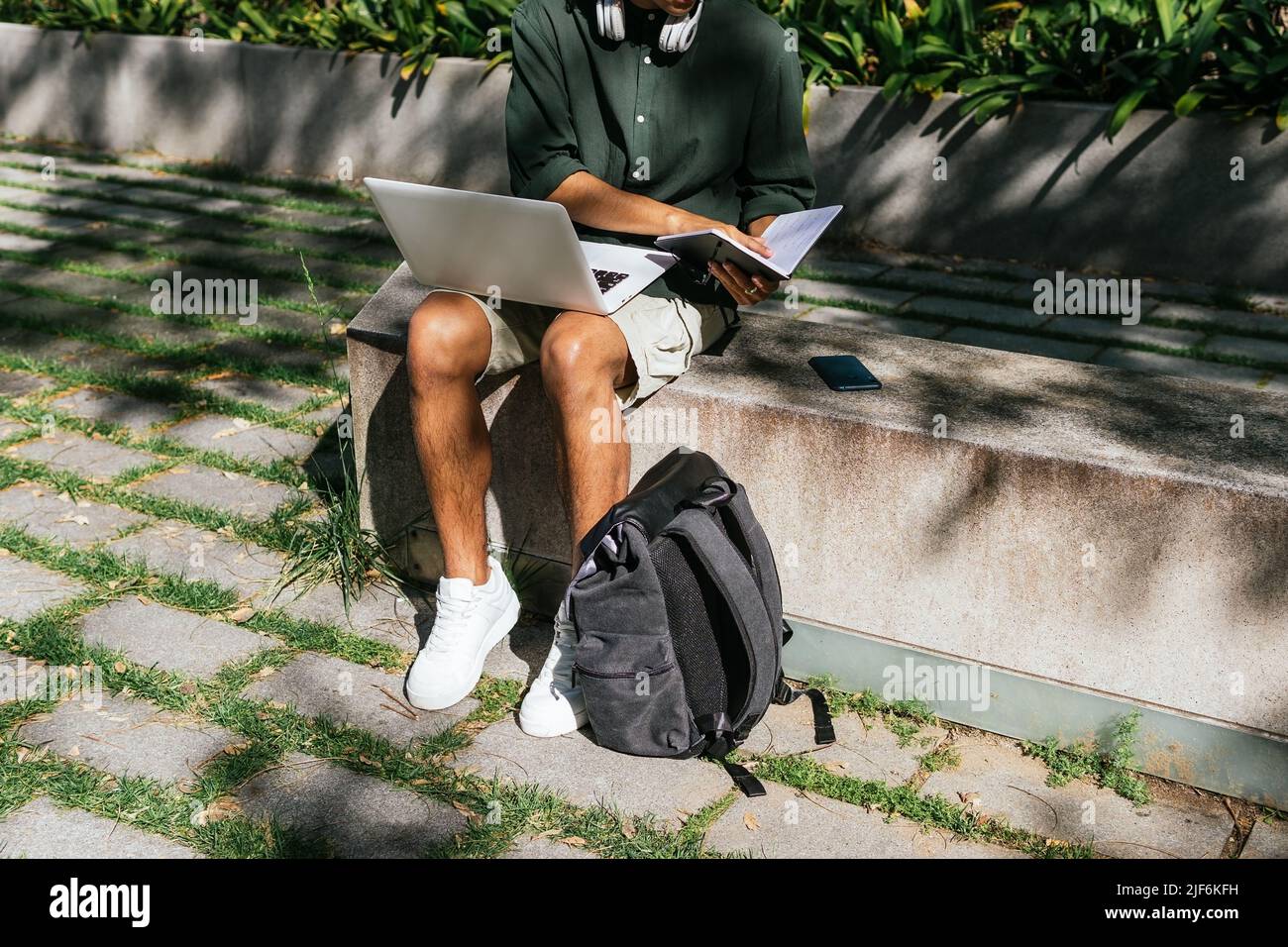 Unrecognizable male student sitting on stone bench in city park and reading notes in notepad while working on assignment using modern netbook Stock Photo