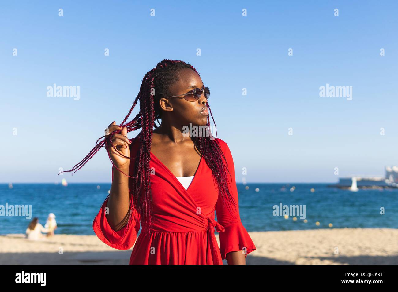 African American young woman in trendy red dress and braids touching hair at the beach Stock Photo