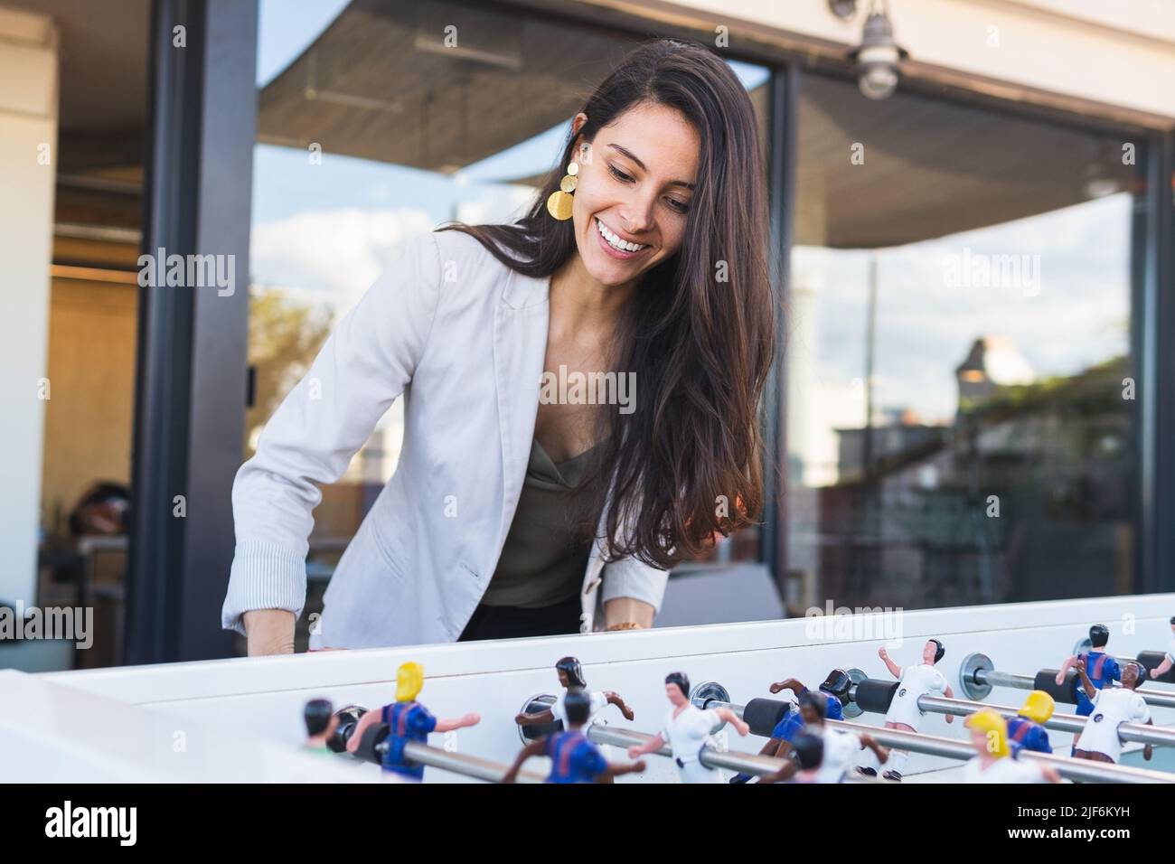 Happy adult Hispanic woman in smart casual clothes smiling and playing table football during party on weekend day on terrace Stock Photo
