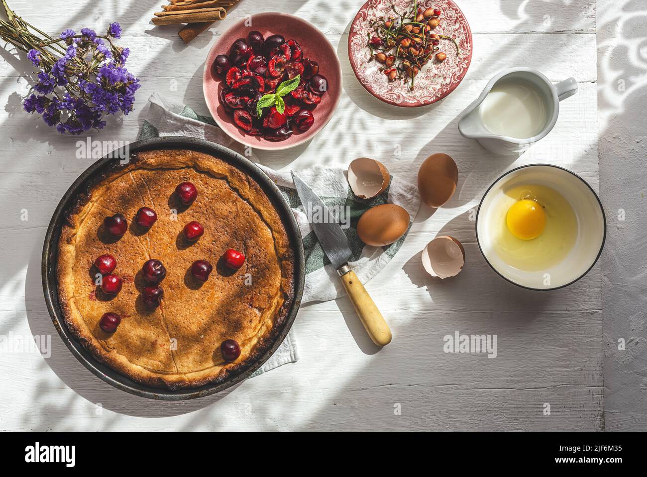 From above baked cherry pie on kitchen cloth near cherry seeds, eggs, shells and jug of milk on white background Stock Photo