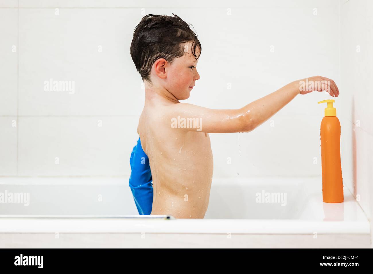 Side view of positive child in waterproof arm plaster protection standing in bathtub and squeezing shampoo while washing hair in white bathroom at hom Stock Photo