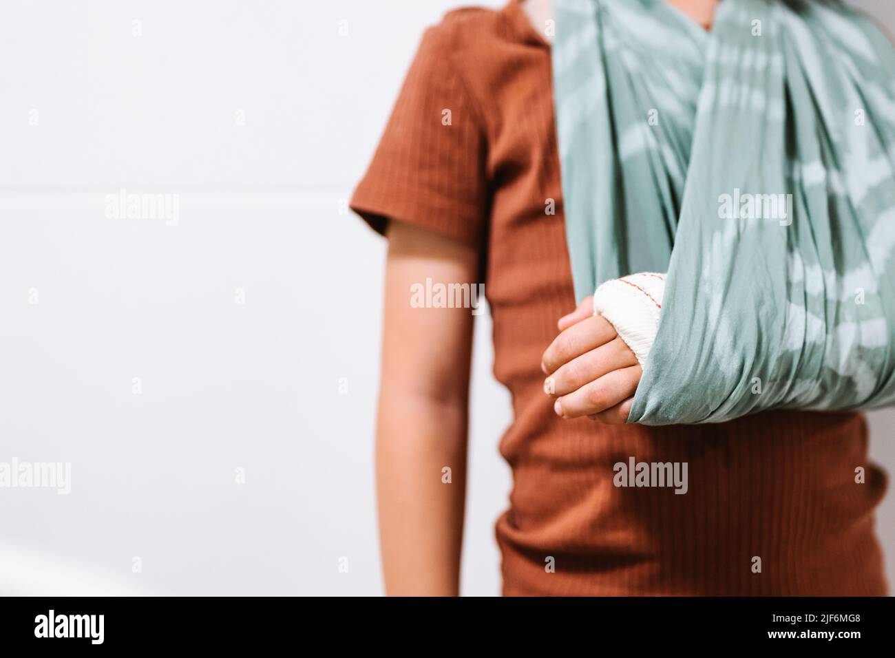 Cropped unrecognizable cute boy touching broken arm in cloth sling standing against white wall Stock Photo