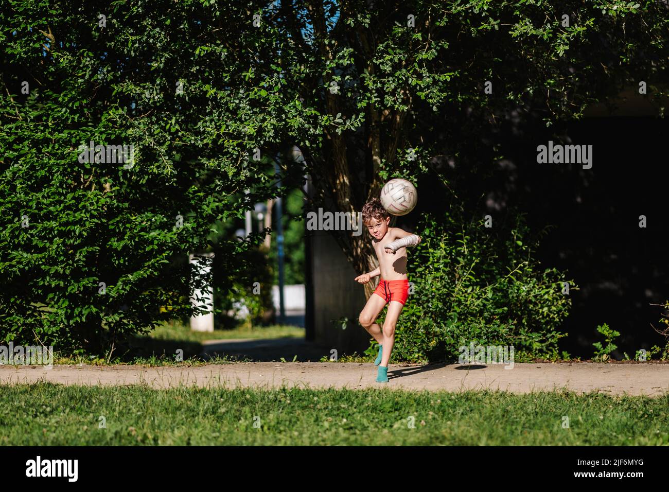 boy in red pants and casted arm kicking soccer ball on green lawn Stock Photo