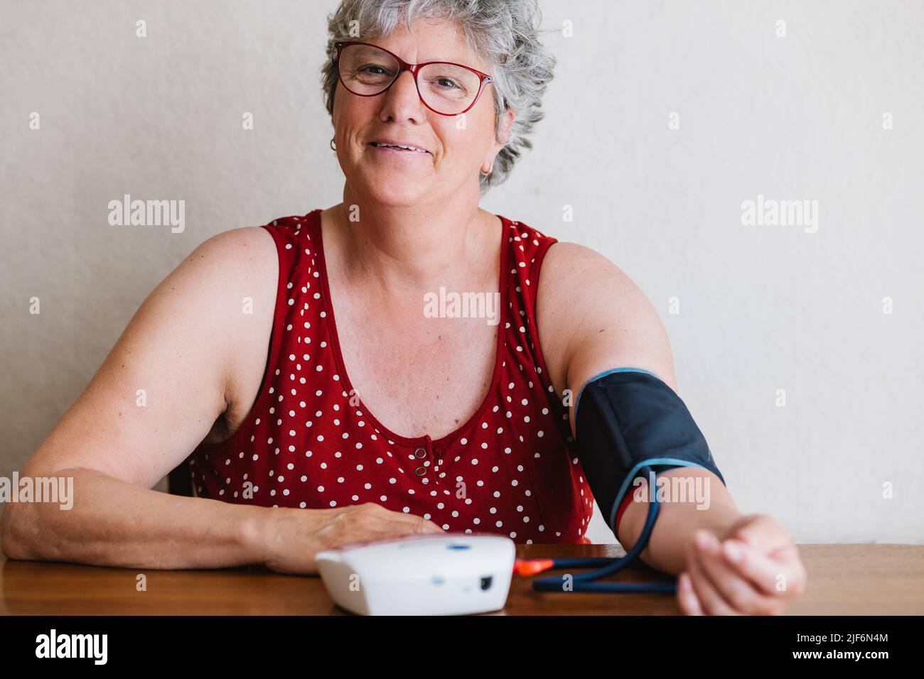 Positive senior female with gray hair in casual clothes and eyeglasses measuring blood pressure with modern sphygmomanometer sitting at table at home Stock Photo