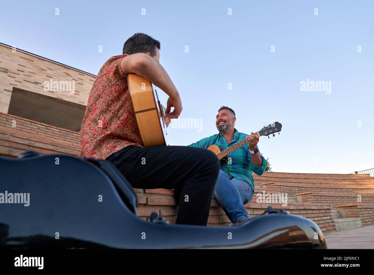 From below positive male musicians playing song on acoustic guitars while sitting together on steps on street of city Stock Photo