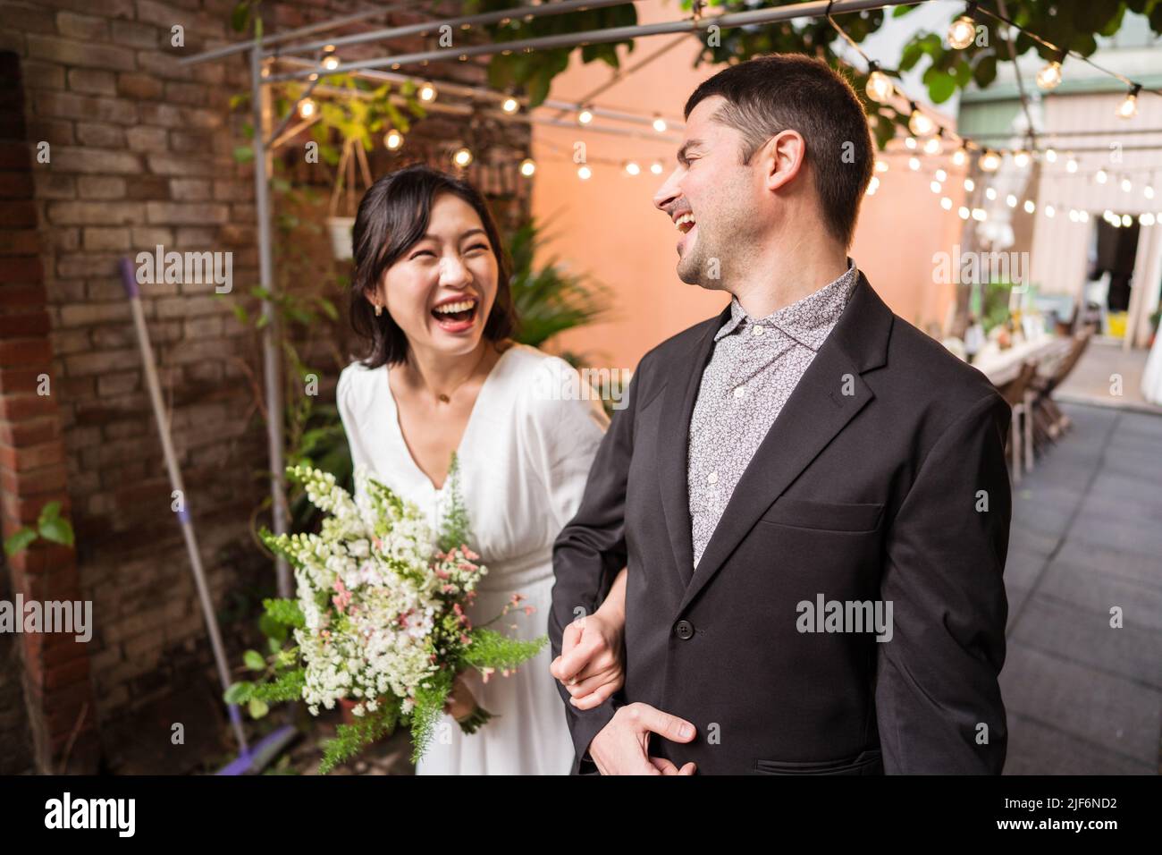 Loving Multiracial couple in wedding outfits smiling and looking at each other while celebrating marriage and standing under tree in park Stock Photo