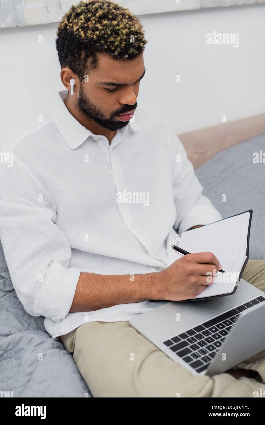 young african american man in wireless earphone making notes near laptop in bedroom Stock Photo