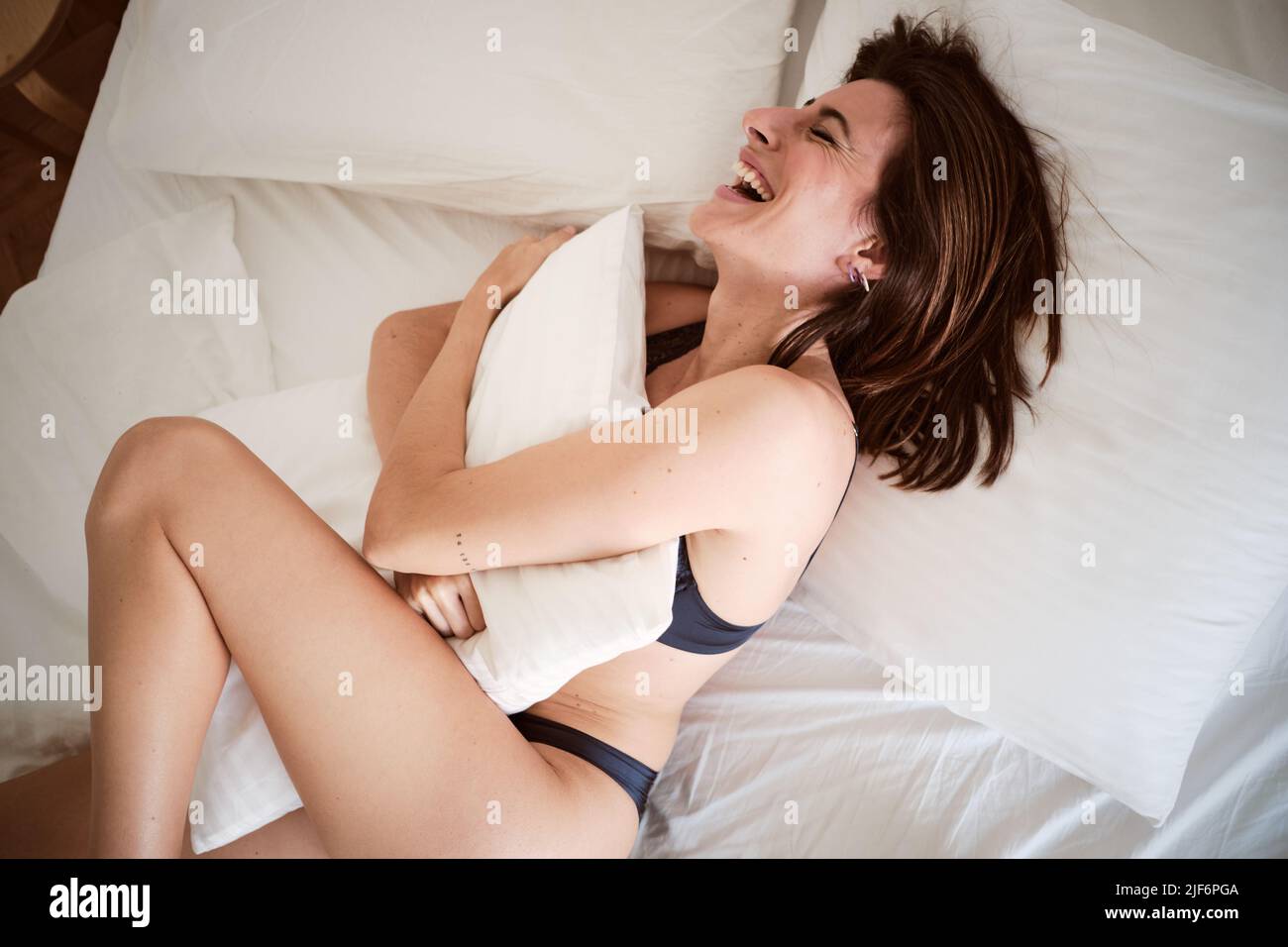 From above of happy female in underwear with eyes closed lying on bed hugging pillow in bedroom Stock Photo