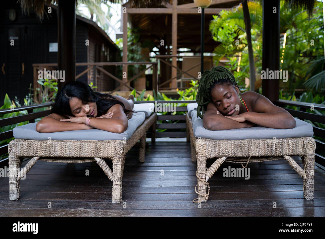 Relaxed black female friends resting on massage tables during spa procedure on terrace of wooden house in tropical country on summer day Stock Photo