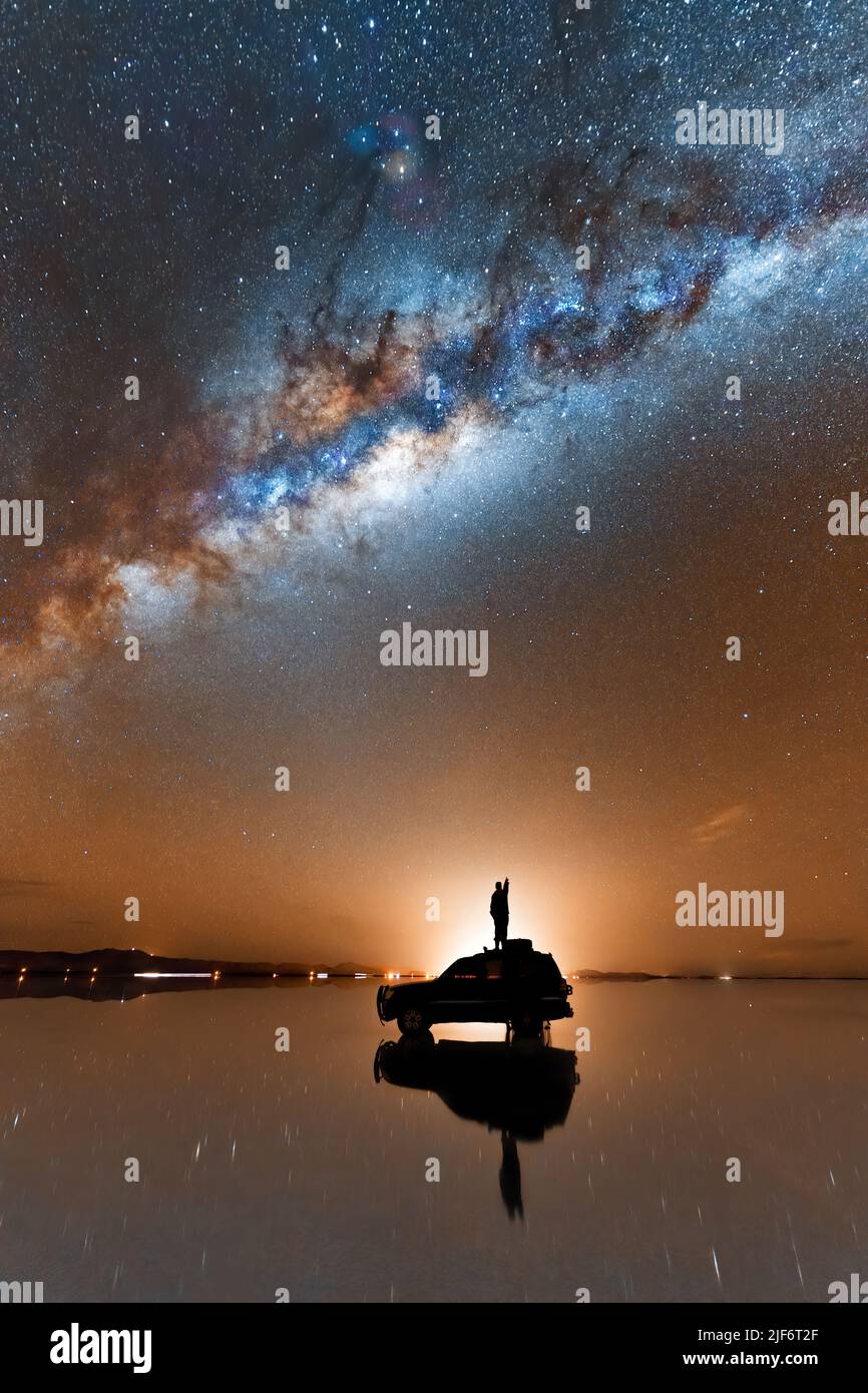 Silhouette of unrecognizable traveler standing on SUV car parked on shiny surface of salt flat of Salar De Uyuni and admiring scenic Milky Way galaxy Stock Photo