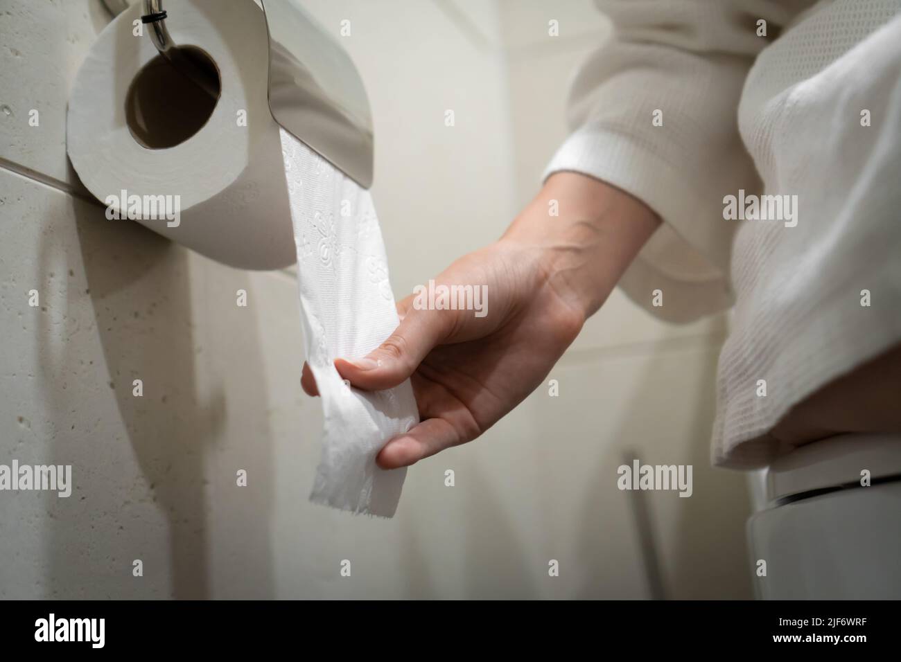 Woman in a white coat sits on the toilet and pulls toilet paper in the bathroom. Selective focus Stock Photo