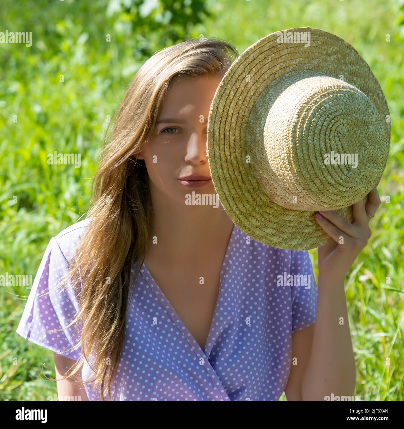 Portrait of a girl in a purple dress covering half of her face with a straw hat and looking at the camera. Stock Photo