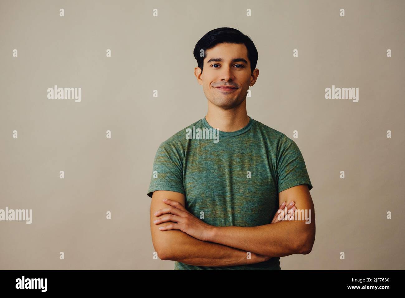 Portrait latino man with arms crossed and black hair smiling handsome young adult green t-shirt over gray background looking at camera studio shot Stock Photo