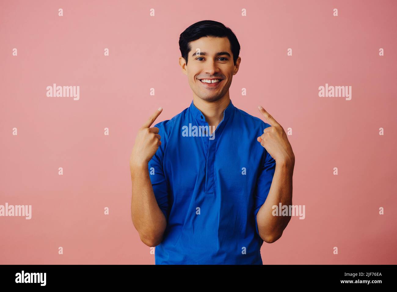 Portrait hispanic latino man with arms crossed pointing at himself black hair smiling handsome young adult blue shirt over pink background looking at camera studio shot Stock Photo