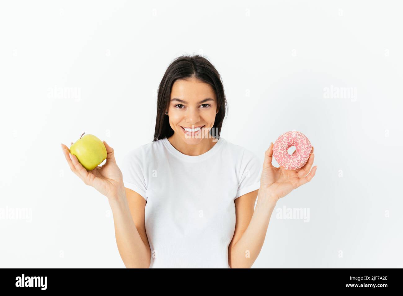 Concept of healthy and unhealthy eating. Happy young woman holding green apple and donut standing against the white studio background Stock Photo