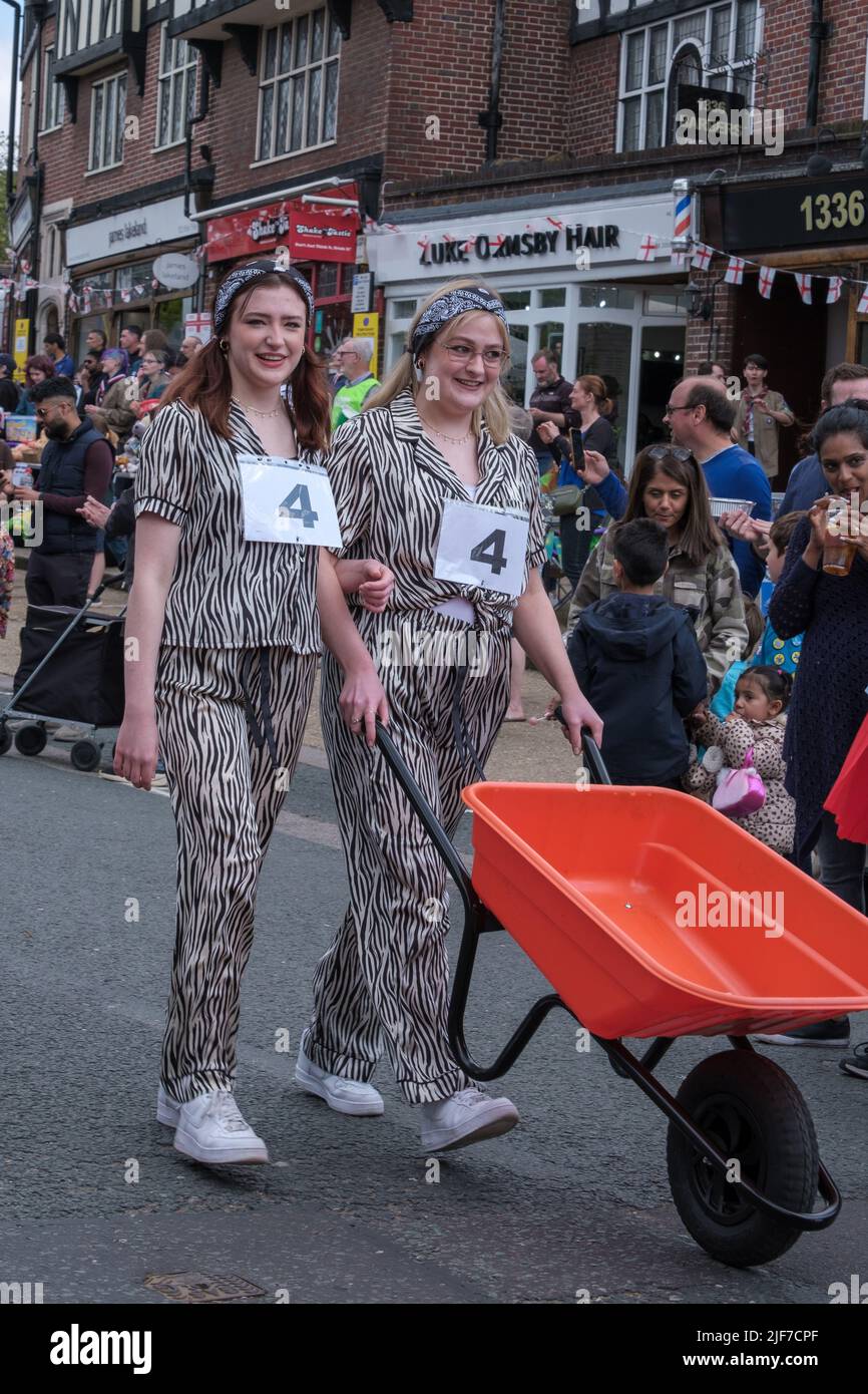 Two ladies in fancy dress push a wheel barrow down Pinner High Street before the wheelbarrow race at St Georges’ Day Celebration. Pinner, NW London. Stock Photo
