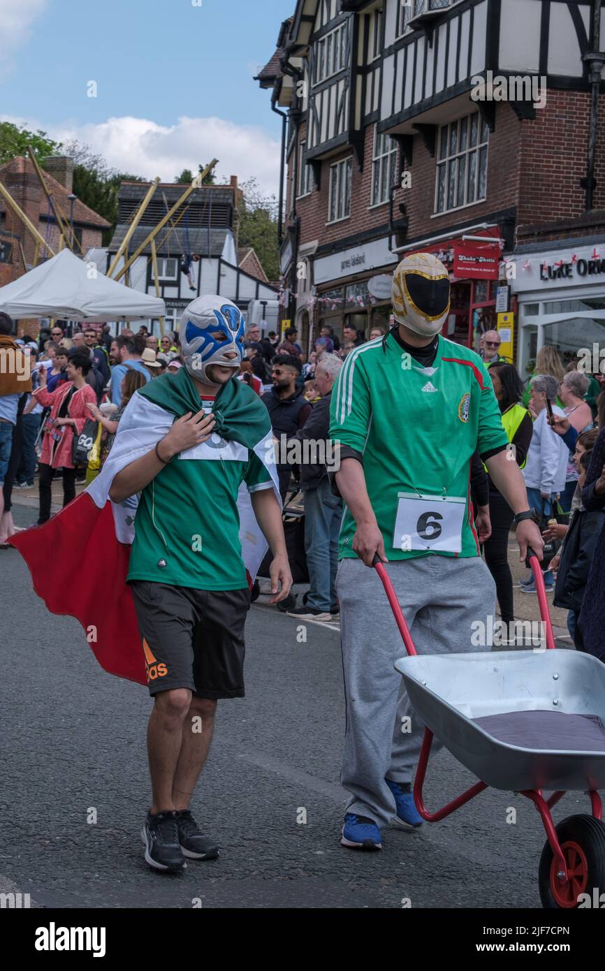 Two men with fancy dress helmets & a cape push a wheel barrow down Pinner High Street before the wheelbarrow race at St Georges’ Day Celebration. Stock Photo