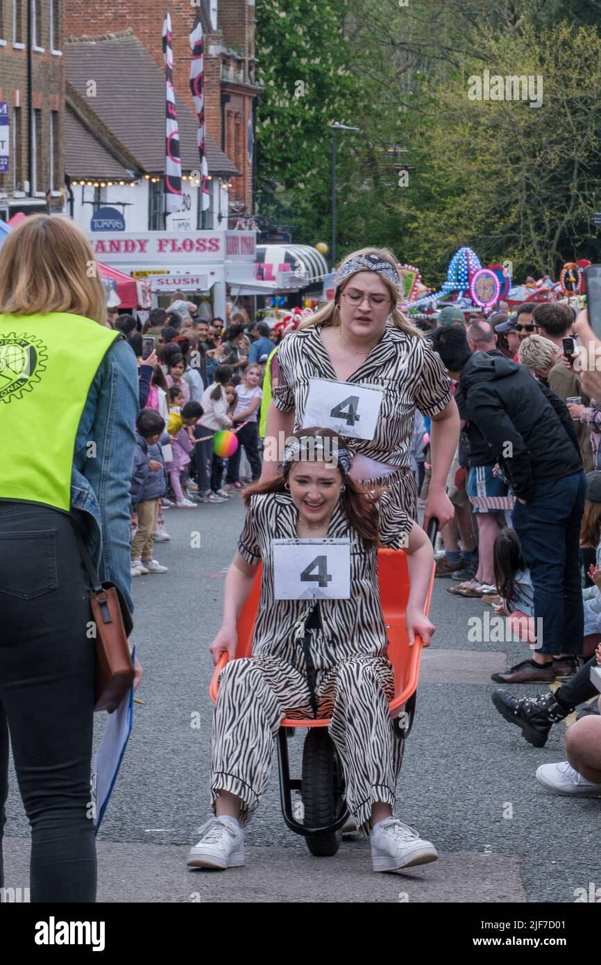 One woman pushes another in a wheelbarrow during the wheel barrow race at St George’s Day Celebration. Pinner, Harrow, North West London. Stock Photo