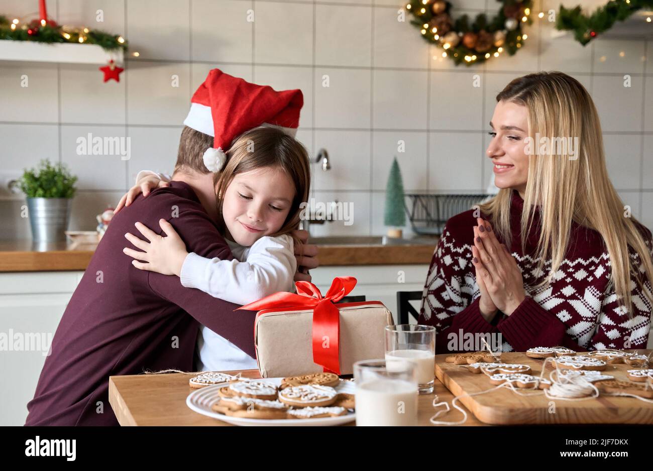 Dad in santa hat presenting Christmas gift to kid daughter hugging at home. Stock Photo