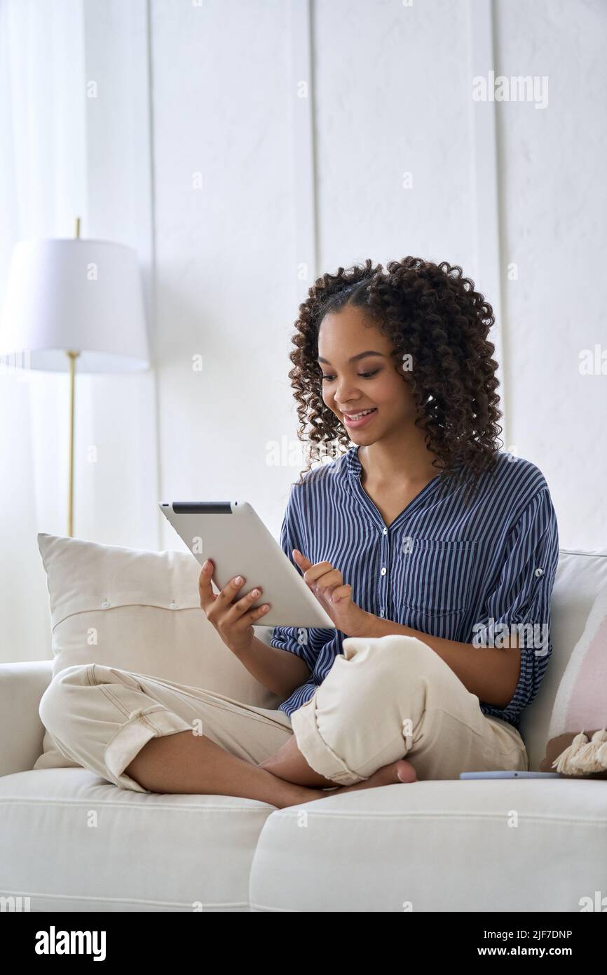 Happy mixed raced African teenage girl sitting on sofa holding using tablet. Stock Photo