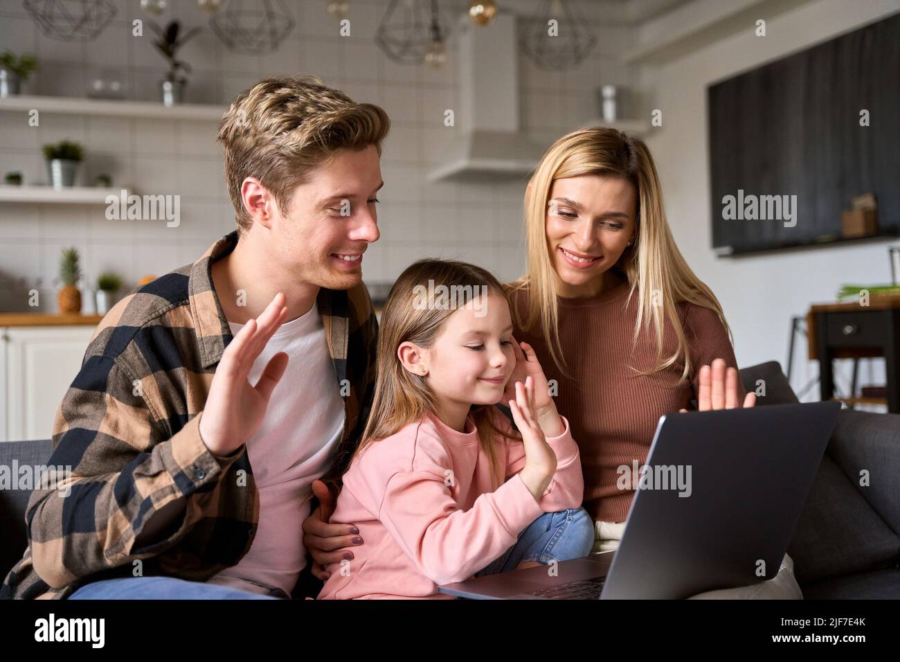 Happy parents with kid daughter waving hands video calling on laptop at home. Stock Photo