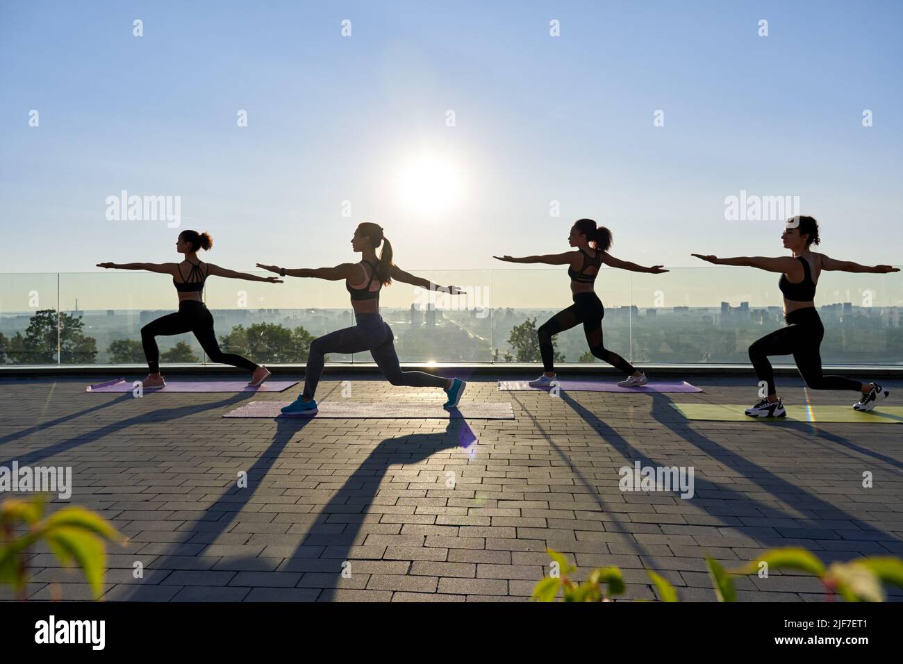 Female silhouettes stand in yoga warrior pose at group class outdoor on sunrise. Stock Photo