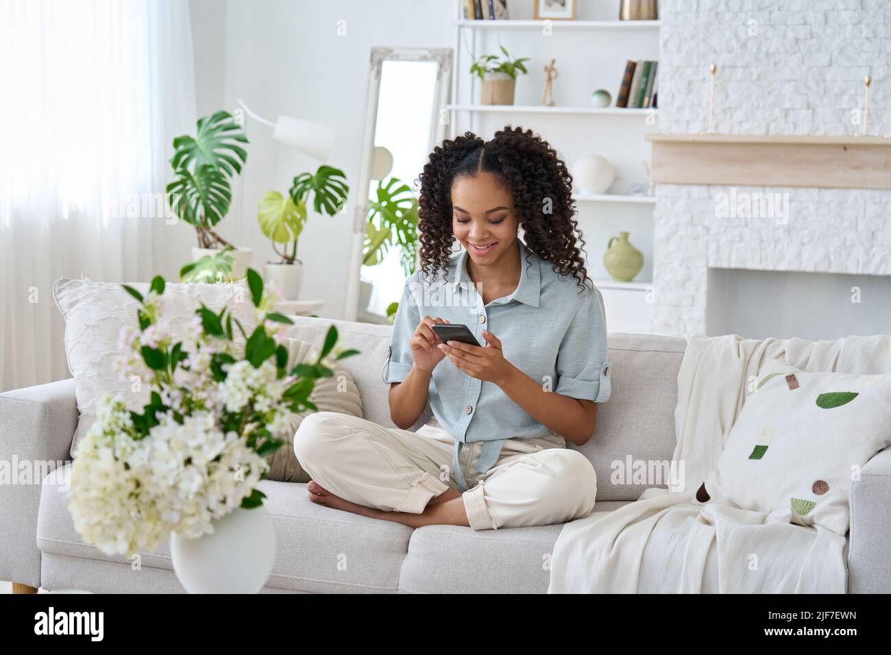 Young happy African woman using mobile apps sitting on sofa at home. Stock Photo