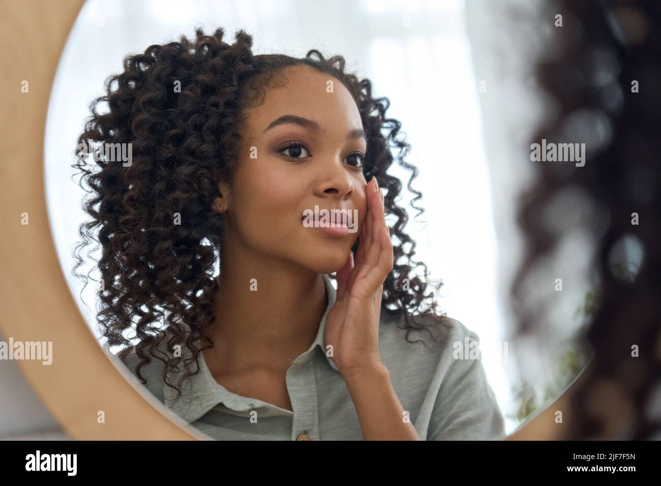 Teen African American girl looking in mirror touching face enjoys healthy skin. Stock Photo