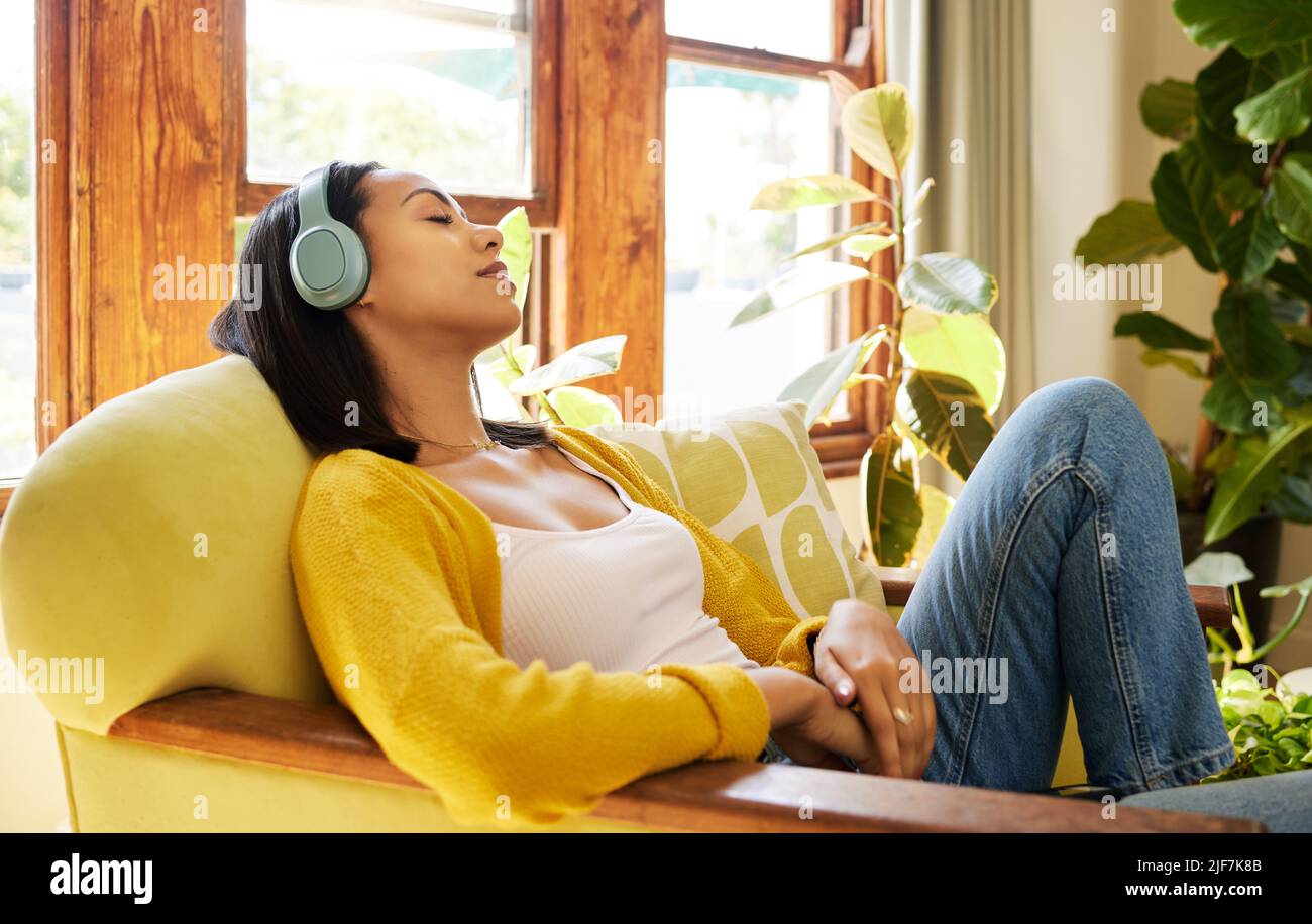 Woman listening to music on headphones while comfortable and relaxing in a bright living room. A young hispanic female with eyes closed resting and Stock Photo